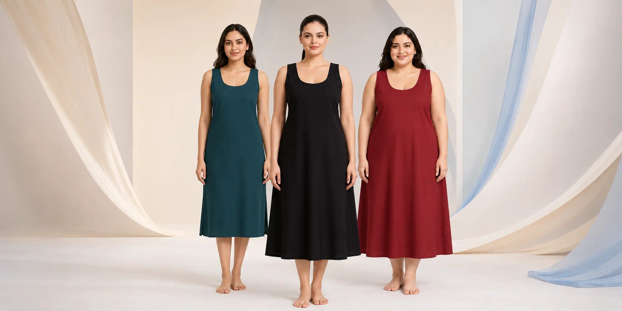 Three women stand barefoot in a studio wearing sleeveless midi-length dress-like garments in three different colors: teal, black, and burgundy. The background is a soft beige and white draped set.
